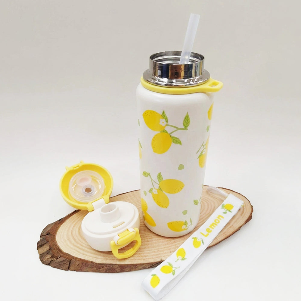 A white insulated water bottle with a yellow lemon print, shown with its lid and straw removed and placed beside it on a round wood slice. A matching wrist strap with lemon illustrations and the word 'Lemon' lies in front.